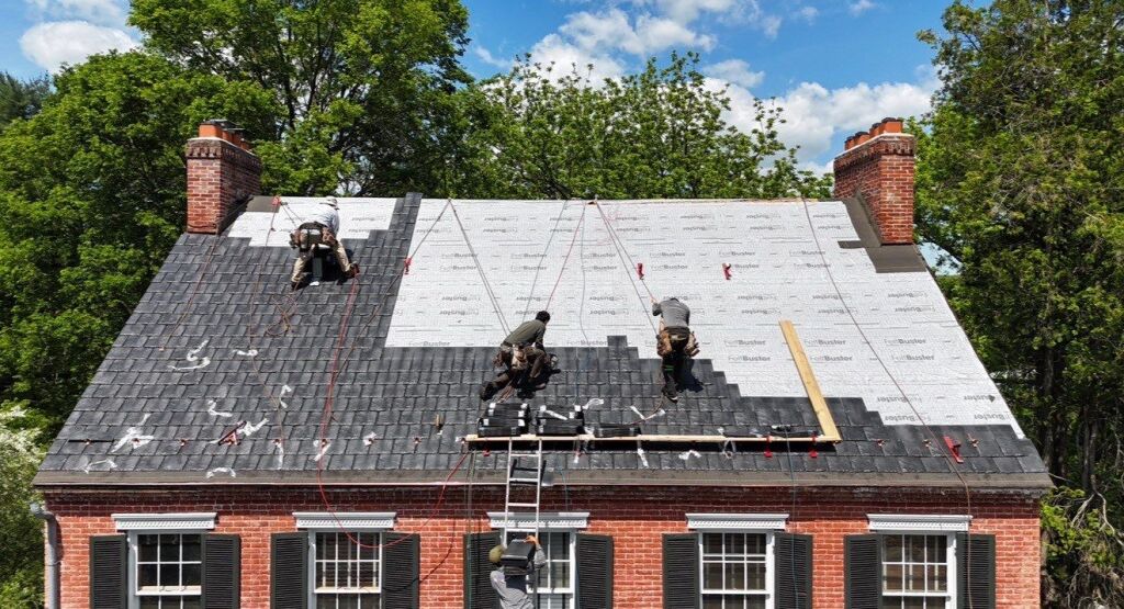 Workers installing shingles on a red brick house roof with safety gear.