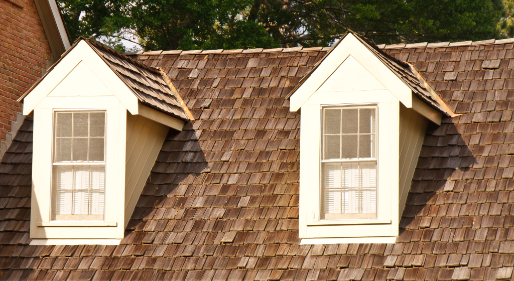 Close-up of shingled roof with two dormer windows and trees in background.